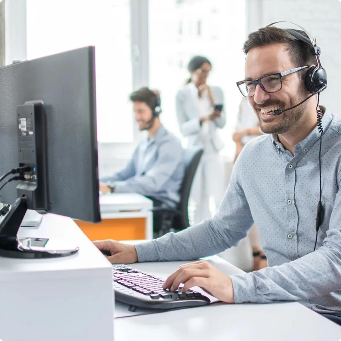 A man wearing a headset and glasses works on a computer, smiling. In the background, two people converse near another person at a computer.
