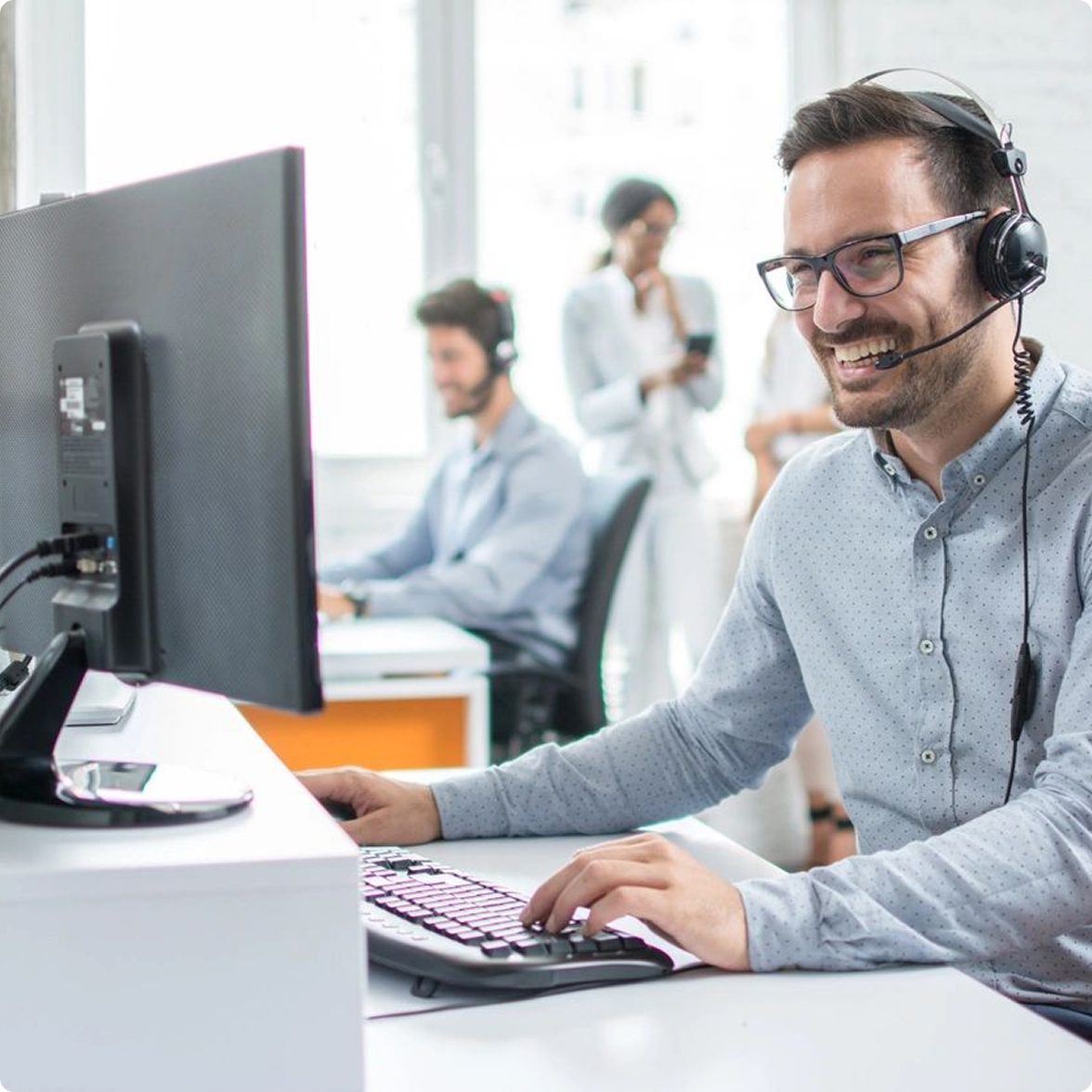 Rectangle 3 (2) A man wearing a headset and glasses works on a computer, smiling. In the background, two people converse near another person at a computer.